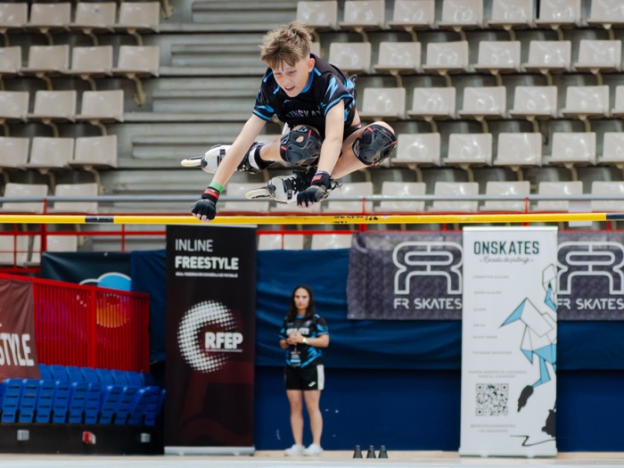 Patinador de Onskates en pleno salto sobre el listón, con las banderolas del club y de la RFEP al fondo